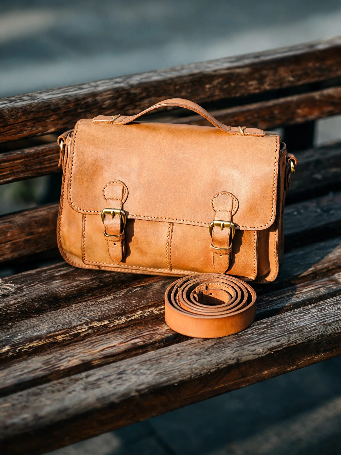 Vegetable-tanned leather satchel with brass buckles beside a coiled belt on a wooden bench
